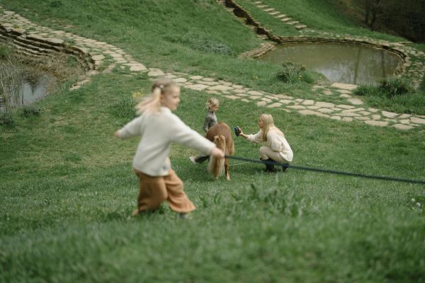 A woman and two children interact with a pony beside a pond in a lush outdoor setting.