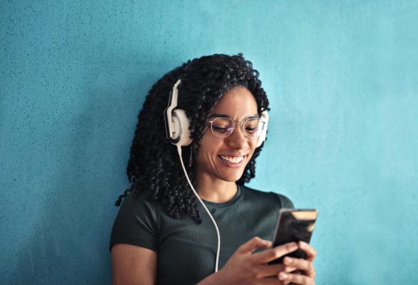 Happy woman with curly hair using smartphone and headphones against teal background.